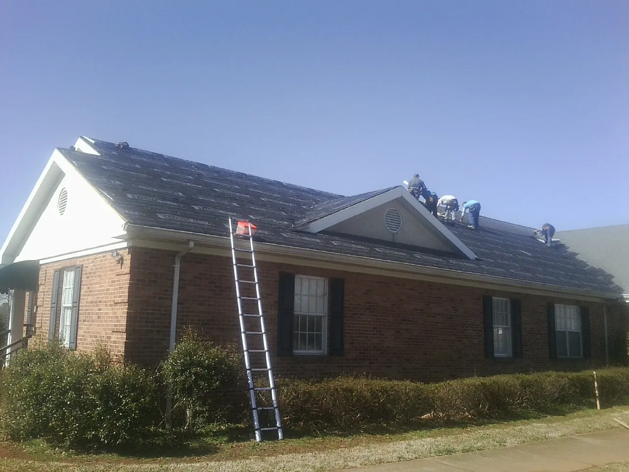 Crew tearing shingles off a brick ranch roof with a ladder set — Emergency Roof Repair work by our crew in Ridgewood