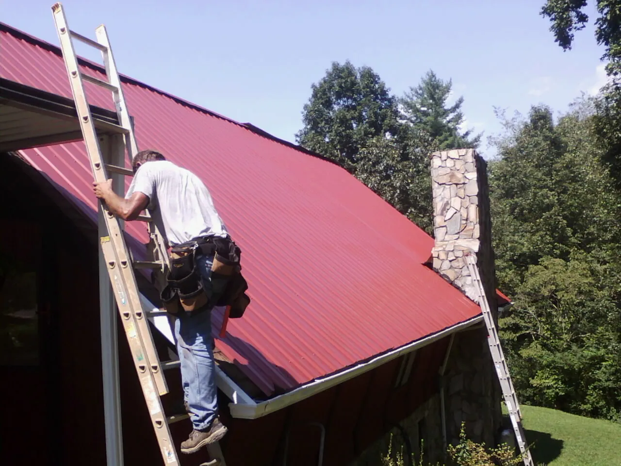 Roofer on a ladder beside a red metal roof and stone chimney — Soffit & Fascia Repair by Roofing Brothers in Ridgewood